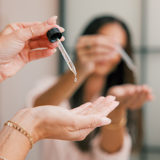 Dropper releasing a drop of skincare serum into an outstretched palm, blurred person and mirror in background