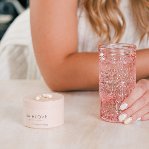 HAIRLOVE growth complex supplement jar with two capsules beside a hand holding an embossed pink glass