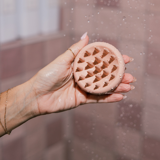Hand holding wet pink silicone scalp-massage brush with short rounded bristles against tiled shower background