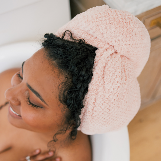 Smiling woman with curly hair wearing a pink textured towel turban while reclining in a bathtub with eyes closed.