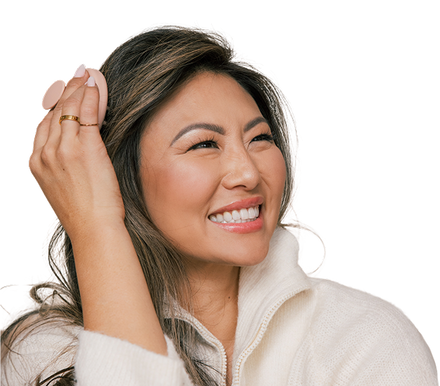 Smiling woman holding a round pink makeup sponge to her temple, wearing a cream zip-up sweater and rings on her fingers.