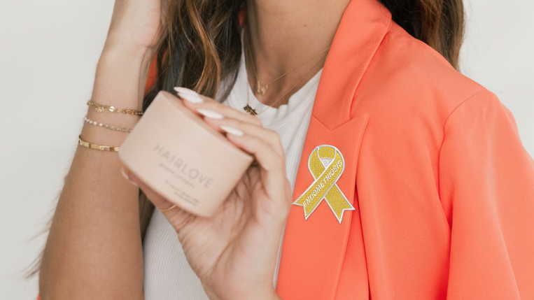 Woman holding a HAIRLOVE jar wearing coral blazer with yellow endometriosis awareness ribbon