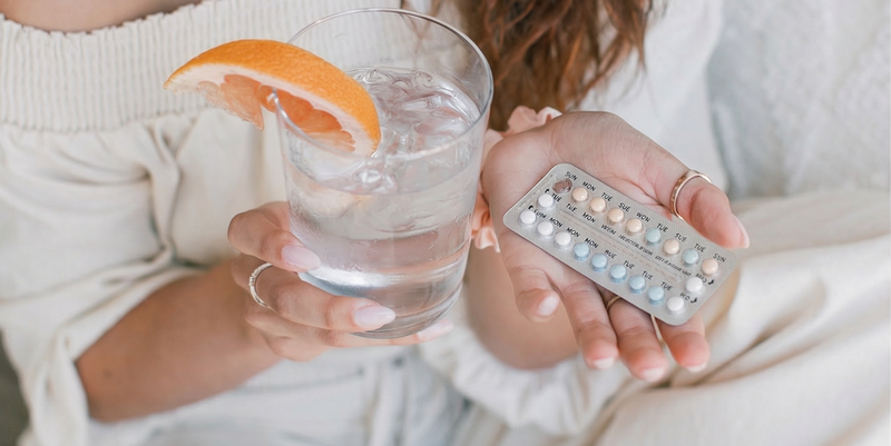 Person holding a blister pack of birth control pills and a glass of water with a grapefruit wedge while sitting on a bed