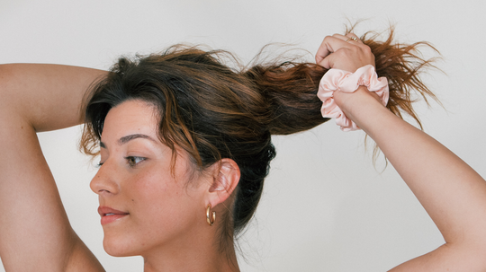 Woman gathering her hair into a ponytail using a pale pink scrunchie against a neutral background