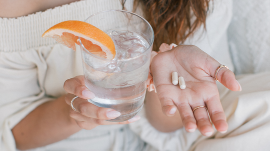 Person holding two supplement capsules in one hand and a glass of water with a grapefruit wedge, seated on a light sofa