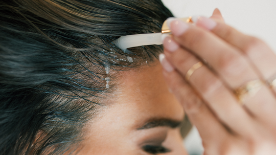 Close up of a woman applying scalp serum with a dropper to her hairline, showing serum droplets and her hand with rings