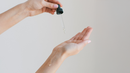 Hands using a dropper to apply argan oil to a palm against a neutral background