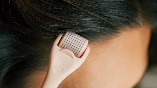 Derma roller rolling across a scalp in a close-up showing the microneedle head and dark hair