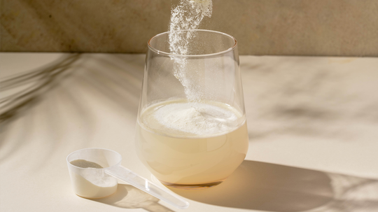 Close-up of creatine powder being poured into a glass showcasing the powder dissolving, with a scoop on a beige tabletop