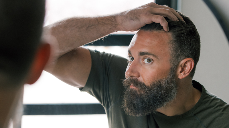 Bearded man examining his hairline in a mirror, inspecting for thinning