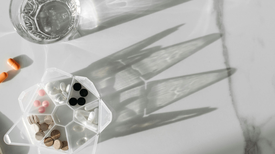 Pill organizer with assorted tablets and capsules beside a glass of water on a marble countertop with long sunlit shadows