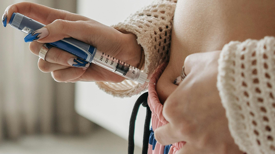 Close-up of a person self-injecting medication into their abdomen with an injector pen while pulling down their waistband
