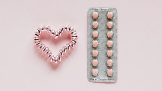 Blister pack of pink pills beside a coiled hair tie shaped into a heart on a pale pink background