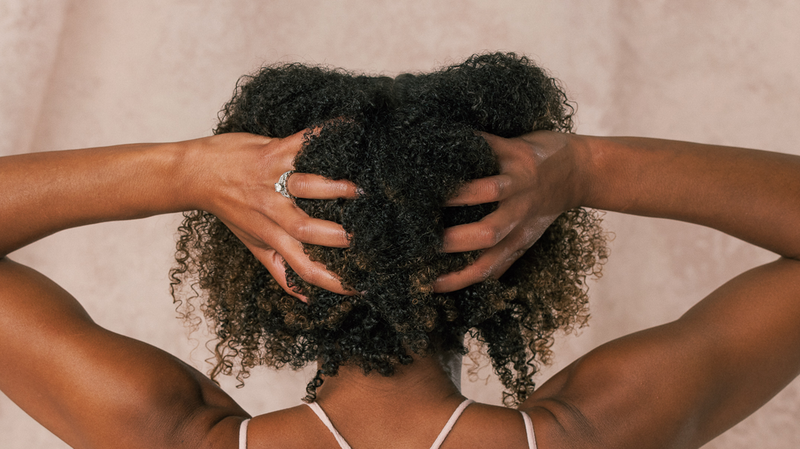 Rear view of a person gathering natural curly hair with both hands against a soft beige background