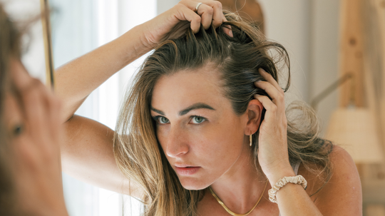 Woman pulling back hair to inspect her hairline in a mirror, appearing concerned in a softly lit bathroom