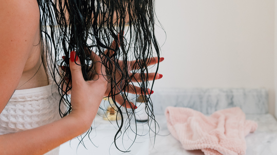 Person with wet dark hair detangling hair over a bathroom sink with a pink towel on the counter