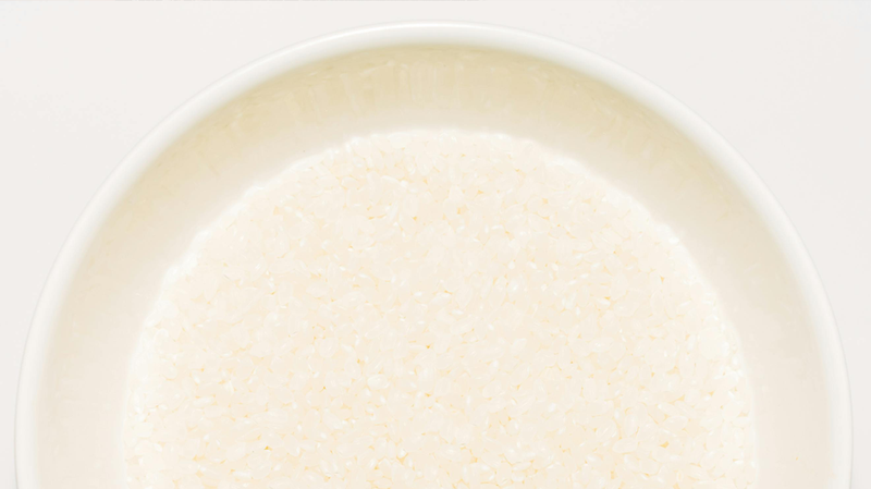 Top-down view of a shallow white bowl filled with uncooked white rice on a pale background