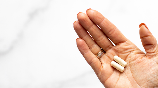 Hand holding two beige supplement capsules against a white marble background, illustrating DHEA supplementation