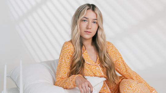 Young woman with long blonde hair sitting on a white bed in yellow floral pajamas, bathed in soft window light
