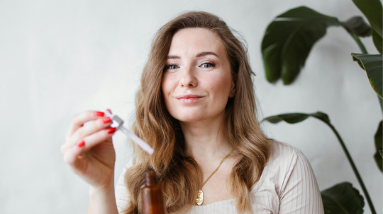 Woman holding a dropper bottle to apply a topical hair treatment in a bright room with a potted plant behind her