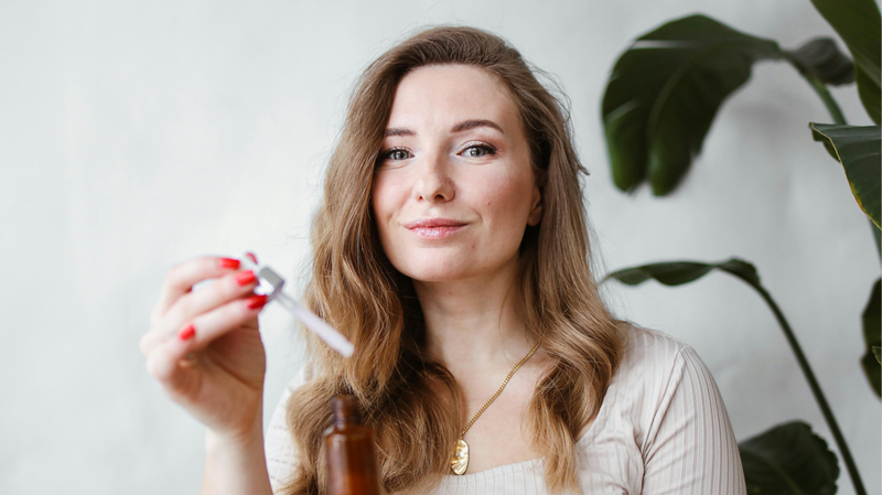 Woman holding a dropper bottle to apply a topical hair treatment in a bright room with a potted plant behind her