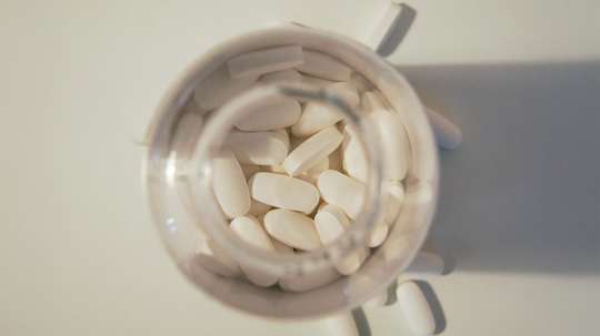 Top-down view of a pill bottle filled with white oblong tablets, several tablets spilled onto a white surface