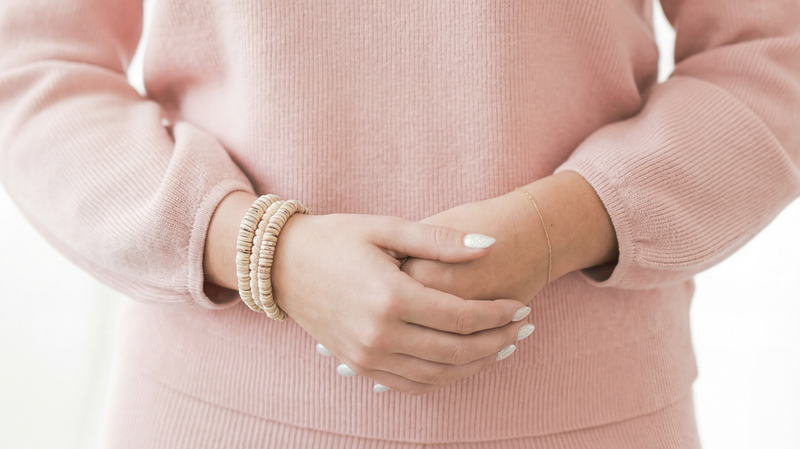 Woman standing with hands clasped in front, wearing a pink ribbed sweater and bracelets against a soft white background