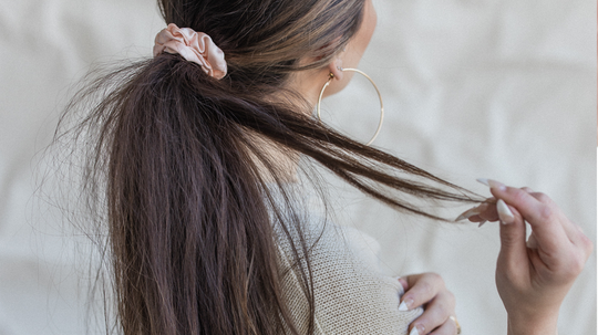 Woman with long brown hair in a low ponytail with a scrunchie holding a loose strand against a neutral background