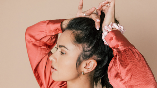 Woman tying her dark hair into a high ponytail wearing a coral satin blouse against a beige background