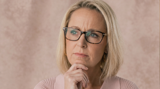 Middle-aged woman with short blonde hair and glasses looking thoughtful against a soft pink background