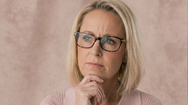 Middle-aged woman with short blonde hair and glasses looking thoughtful against a soft pink background