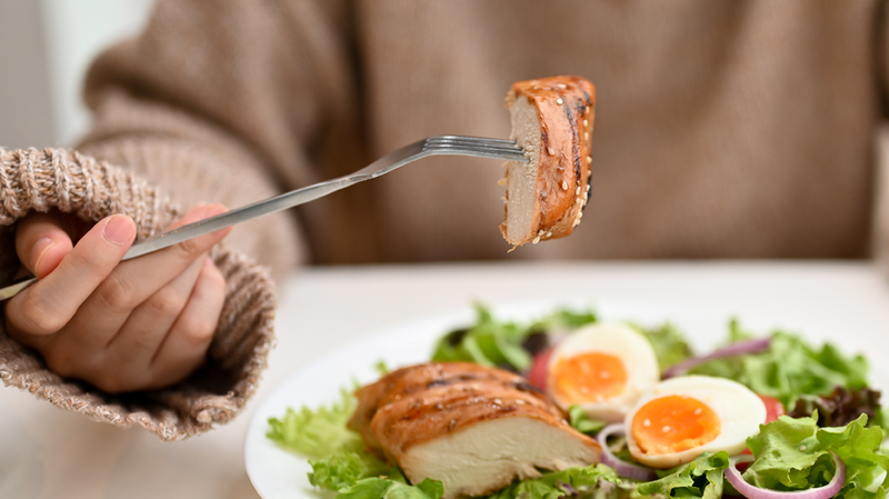 Person holding a forkful of chicken over a salad with soft boiled eggs and greens, suggesting a keto-friendly meal