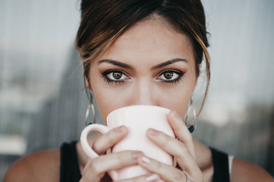 Close-up of woman holding a white mug to her face, looking at camera and highlighting her under-eye area