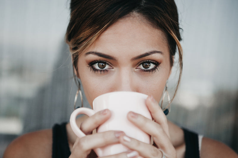 Close-up of woman holding a white mug to her face, looking at camera and highlighting her under-eye area