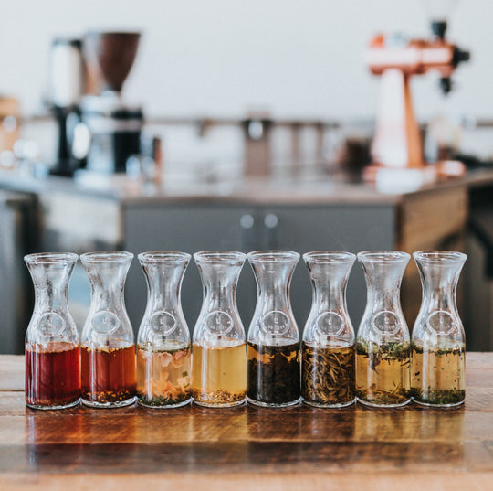 Eight glass carafes of steeping tea lined on a wooden table with a blurred kitchen background