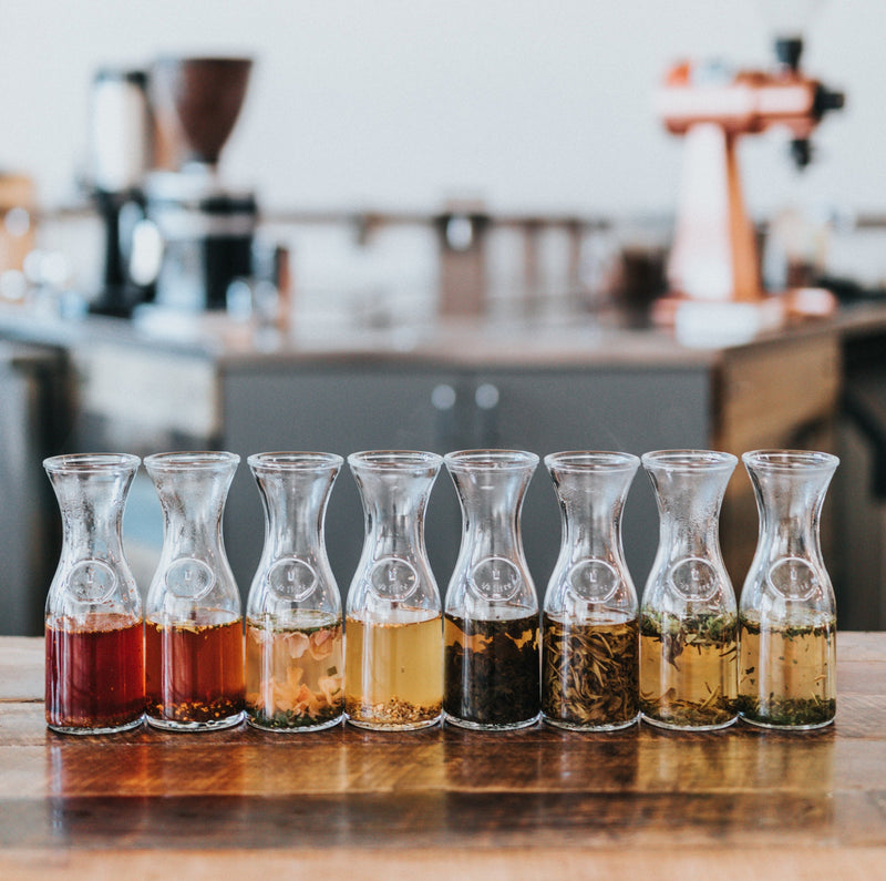 Eight glass carafes of steeping tea lined on a wooden table with a blurred kitchen background