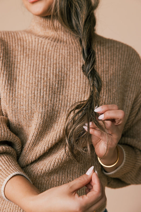 Close-up of a woman in a brown sweater holding and examining the ends of her long braided hair, showing split ends.