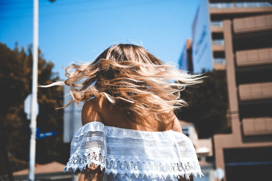 Woman flipping sunlit shoulder-length highlighted hair on a sunny city street, wearing a white off-shoulder lace top