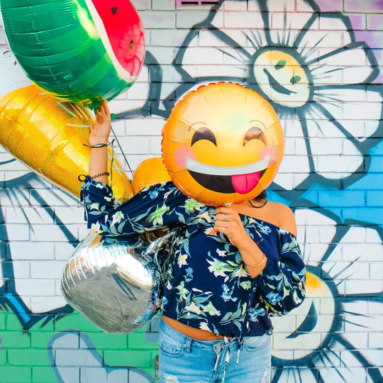 Person holding colorful emoji balloons, including a smiling face with tongue out, in front of a graffiti wall with a smiling flower, conveying happiness and positivity.