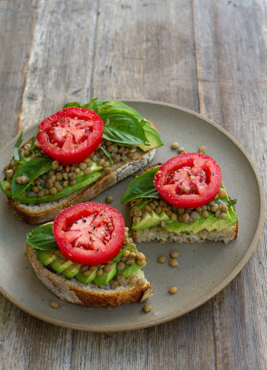 Three slices of avocado toast topped with lentils, basil, and tomato on a ceramic plate over a wooden table