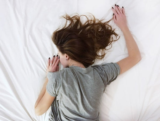 Woman resting face down on a white bed with hair fanned out, wearing a gray t-shirt and dark nail polish, illustrating a nighttime beauty ritual