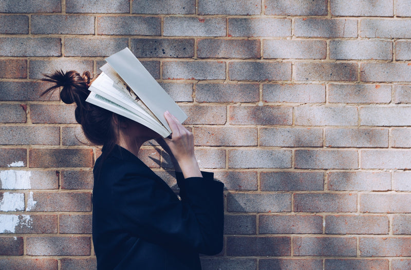Person with hair in a bun holding an open book over their face while standing against a sunlit brick wall