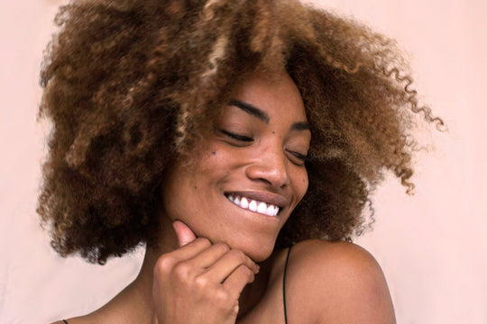 Smiling woman with natural curly hair touching her chin against a pale pink background