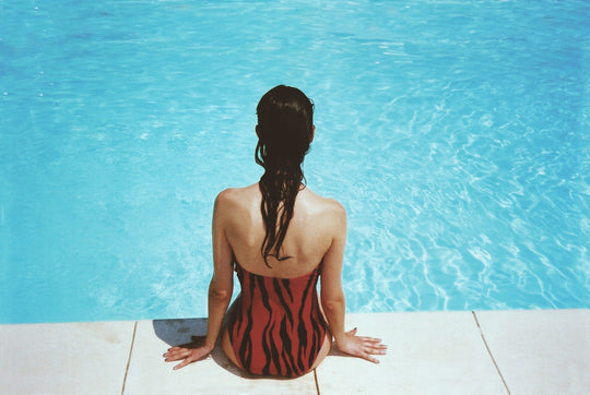 Woman with wet hair sitting on a pool edge facing bright blue water, wearing a red and black swimsuit