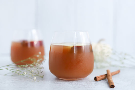 Two glasses of apple cider vinegar drink on a light marble surface, garnished with a lemon slice, cinnamon sticks, and small white flowers