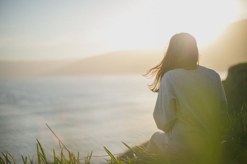 A woman sitting on grass by the ocean, looking at the sunset