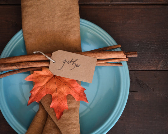 Place setting with blue plate, brown napkin, cinnamon sticks, and a tag reading gather on a dark wood table