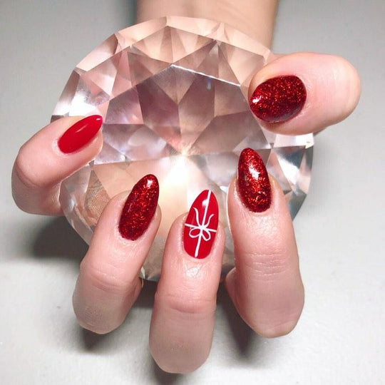 Close up of hand with red almond shaped nails, glitter accents and white bow nail art holding a faceted clear crystal