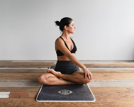 Woman in black sports bra and shorts seated cross-legged on a yoga mat, performing a seated spinal twist pose in a bright room with wooden floor and white wall.