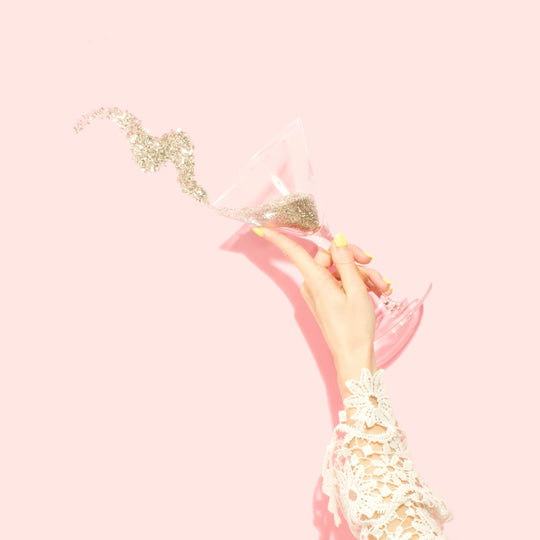A hand holding a champagne flute with sparkling glitter spilling out against a pink background.